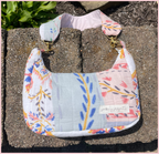 A handmade quilted shoulder bag with a floral patchwork print in pastel colors, resting on concrete blocks outdoors. The bag features a light blue-green center panel, pink and white floral sections, and gold-tone hardware.