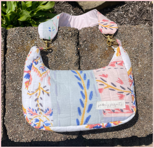 A handmade quilted shoulder bag with a floral patchwork print in pastel colors, resting on concrete blocks outdoors. The bag features a light blue-green center panel, pink and white floral sections, and gold-tone hardware.