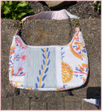 A quilted shoulder bag with a white base and a colorful floral and botanical print in orange, blue, and pink, resting on a stone surface outdoors.