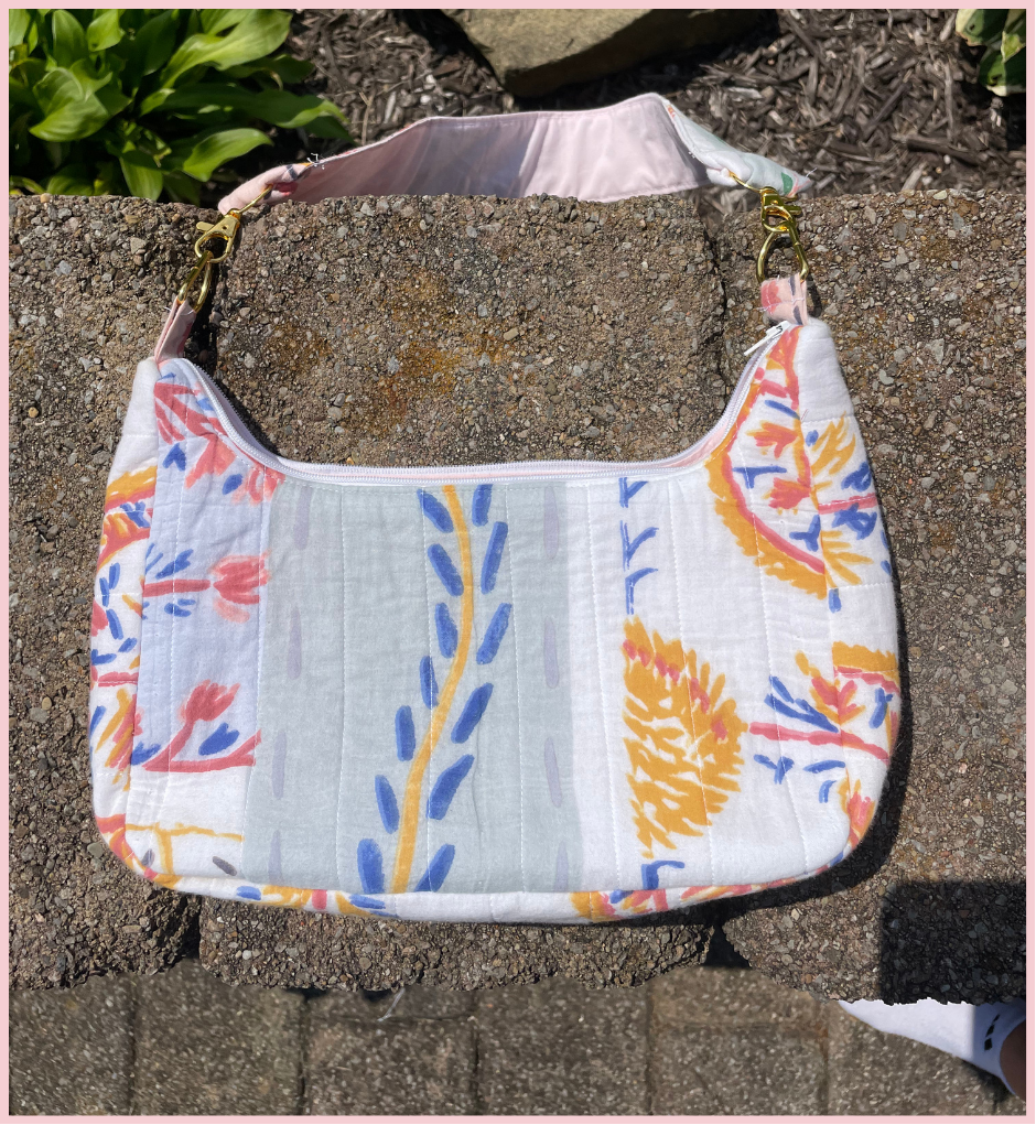 A quilted shoulder bag with a white base and a colorful floral and botanical print in orange, blue, and pink, resting on a stone surface outdoors.