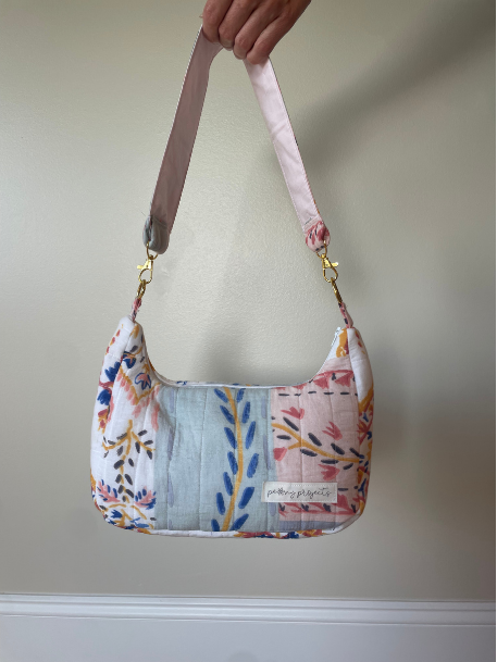 A handmade quilted shoulder bag with a floral and botanical patchwork pattern, held by a hand against a plain beige wall. The bag features a light blue striped center panel, pink and white floral sections, and a dusty pink strap with gold-tone hardware.