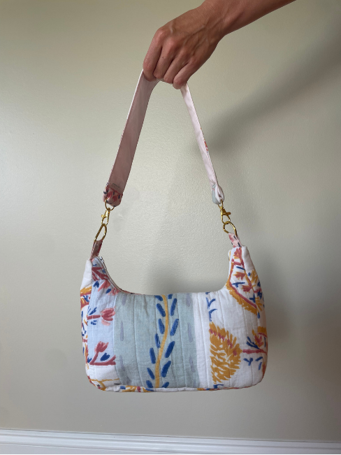 A hand holding a small shoulder bag with a floral and botanical print. The bag features a white base with blue, orange, and red patterns, a light blue center panel, and a light pink adjustable shoulder strap with gold-tone hardware.