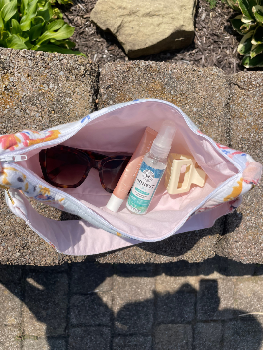A pastel pink zippered travel bag with a colorful floral print on the exterior, shown open on a stone ledge outdoors. Inside, the bag contains sunglasses, a pink tube, a small spray bottle, and a beige hair clip.