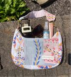 A handmade patchwork shoulder bag with a floral and botanical print, styled outdoors on a stone surface with sunglasses, a hand sanitizer bottle, and a pink tube.