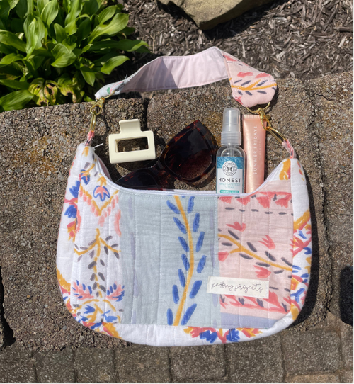 A handmade patchwork shoulder bag with a floral and botanical print, styled outdoors on a stone surface with sunglasses, a hand sanitizer bottle, and a pink tube.
