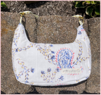 A white quilted shoulder bag with a blue, pink, and yellow botanical pattern, resting on a stone wall outdoors.