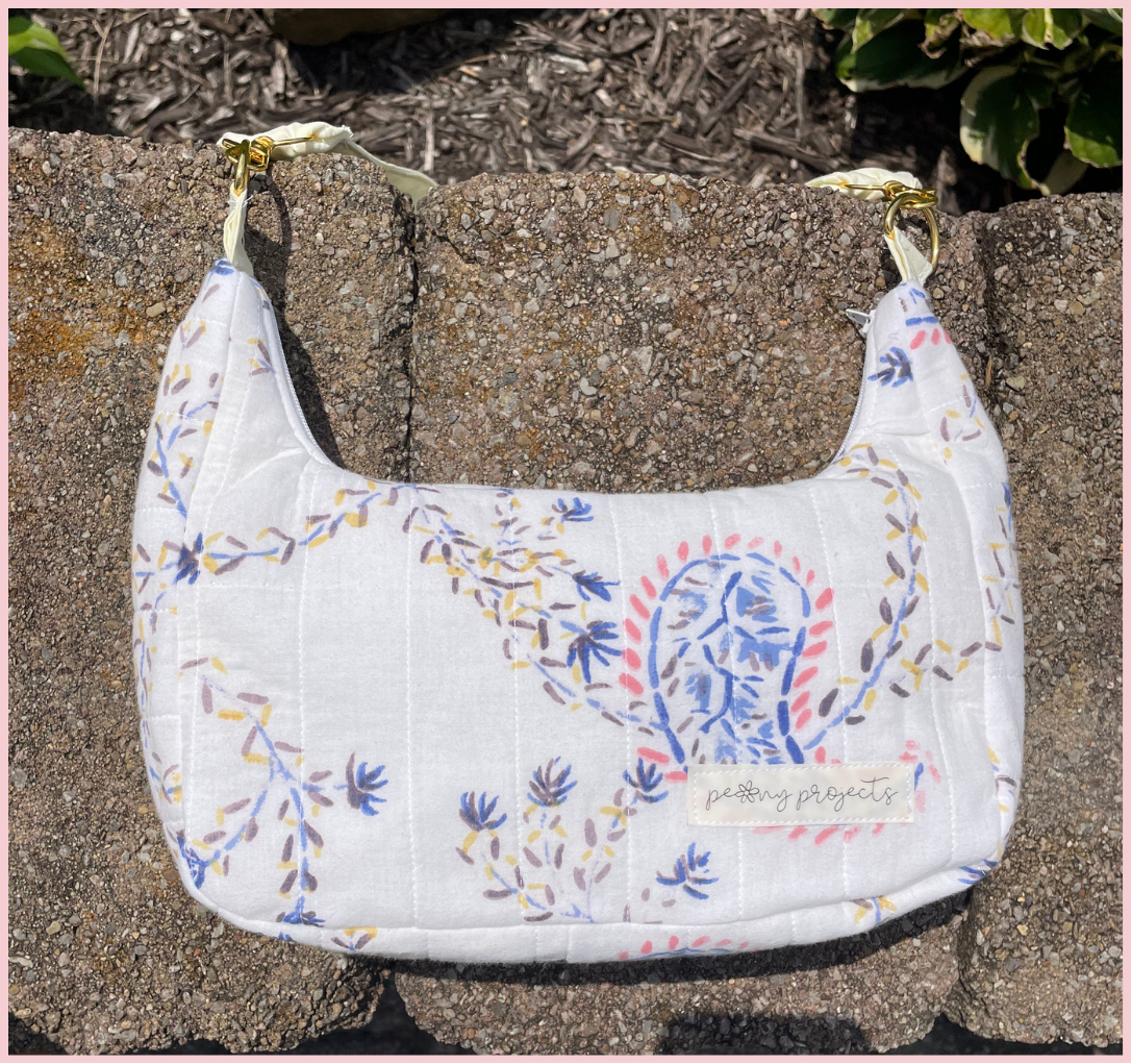 A white quilted shoulder bag with a blue, pink, and yellow botanical pattern, resting on a stone wall outdoors.