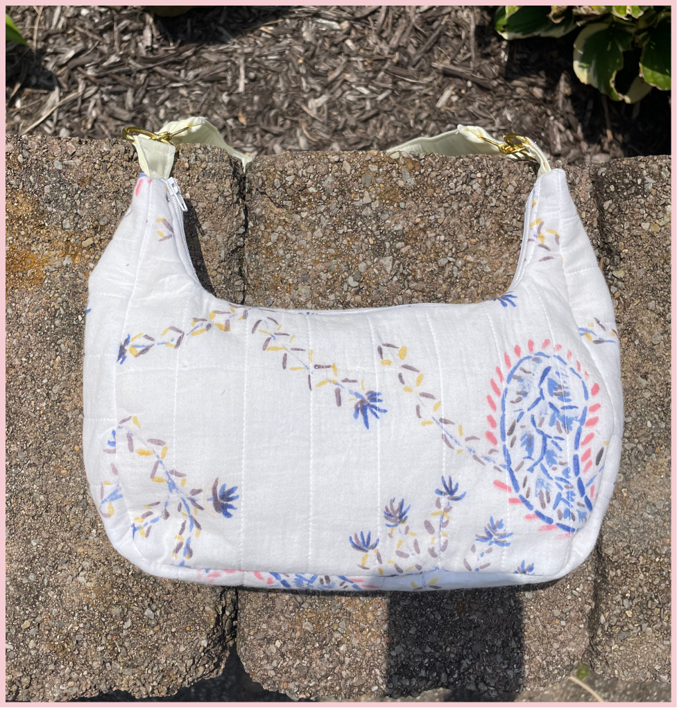 A white quilted fabric shoulder bag with blue, yellow, and red embroidered floral and paisley patterns, resting on a stone surface outdoors.