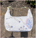 A white quilted fabric shoulder bag with blue, yellow, and red embroidered floral and paisley patterns, resting on a stone surface outdoors.