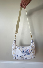 A hand holding up a small quilted shoulder bag with a cream strap and blue floral embroidery on a white fabric, set against a plain beige wall.