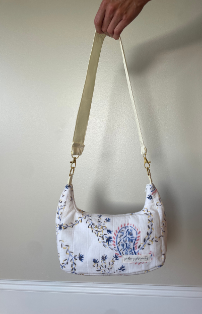 A hand holding up a small quilted shoulder bag with a cream strap and blue floral embroidery on a white fabric, set against a plain beige wall.