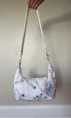 A white shoulder bag with blue and pink floral embroidery, held by a hand against a beige wall.
