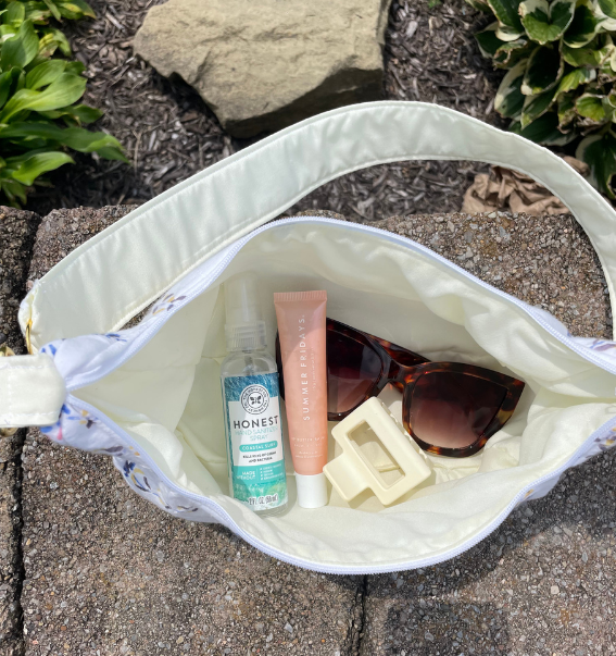 An open white shoulder bag with a floral patterned strap, shown outdoors on a stone surface. Inside the bag are a clear bottle labeled Honest Aesthetic, a pink tube labeled The Ordinary, tortoiseshell sunglasses, and a cream hair clip.