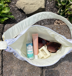 An open white shoulder bag with a floral patterned strap, shown outdoors on a stone surface. Inside the bag are a clear bottle labeled Honest Aesthetic, a pink tube labeled The Ordinary, tortoiseshell sunglasses, and a cream hair clip.