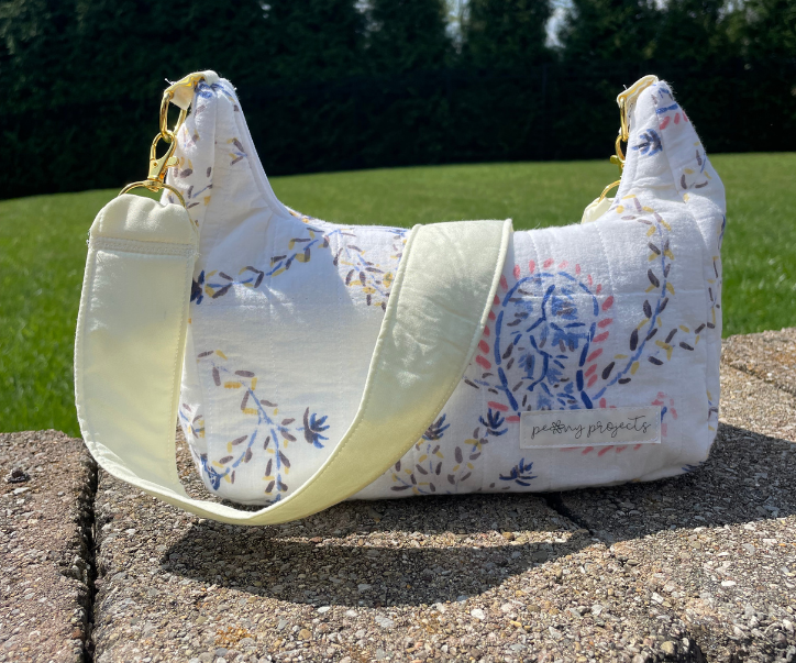 A fabric shoulder bag with a white base and a blue, yellow, and pink botanical pattern, featuring a wide pale yellow-green strap, resting on a stone surface with a green lawn in the background.