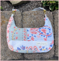 A handmade shoulder bag made from patchwork quilted cotton muslin fabric with a floral print, resting on concrete pavers outdoors. The bag features a light blue, pink, and white pattern with gold-tone hardware and a 'patching projects' brand label.