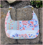 A handmade shoulder bag made from patchwork quilted cotton muslin fabric with a floral print, resting on concrete pavers outdoors. The bag features a light blue, pink, and white pattern with gold-tone hardware and a 'patching projects' brand label.