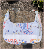 A quilted fabric shoulder bag with a floral and paisley print, resting on a concrete surface outdoors.