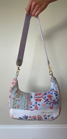 A hand holds up a small shoulder bag with a floral patchwork print in blue, red, and green on a white background. The bag has a wide adjustable shoulder strap in a muted purple-gray color and gold-tone hardware.