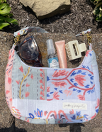 A floral patchwork shoulder bag with a light blue, pink, and red pattern, shown outdoors on a stone surface with sunglasses, a spray bottle, a lip balm tube, and a hair clip inside.