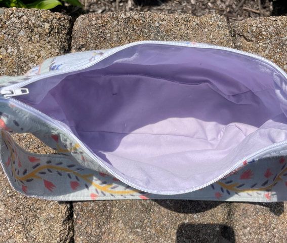An open zippered pouch or bag with a light lavender interior and a floral patterned exterior, photographed outdoors on stone pavers.
