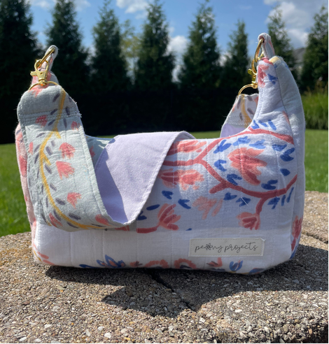 A fabric shoulder bag with a floral pattern in pink, blue, and yellow on a white background, sitting on a concrete surface outdoors with grass and trees in the background. A brand label reading 'pursuing projects' is visible on the front.
