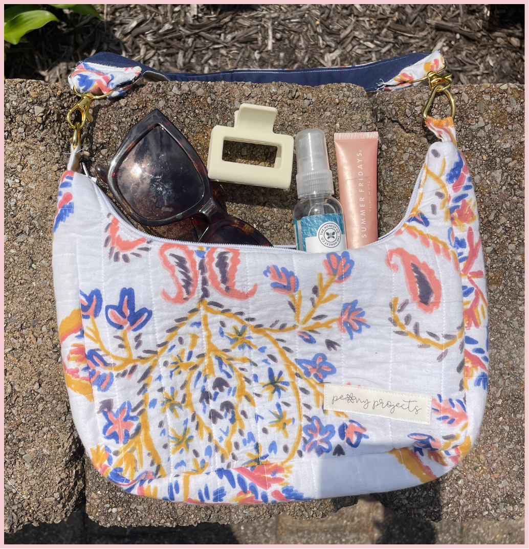 A white shoulder bag with a colorful paisley and floral print, styled outdoors on a stone surface with sunglasses, a hair clip, a spray bottle, and a pink tube inside.