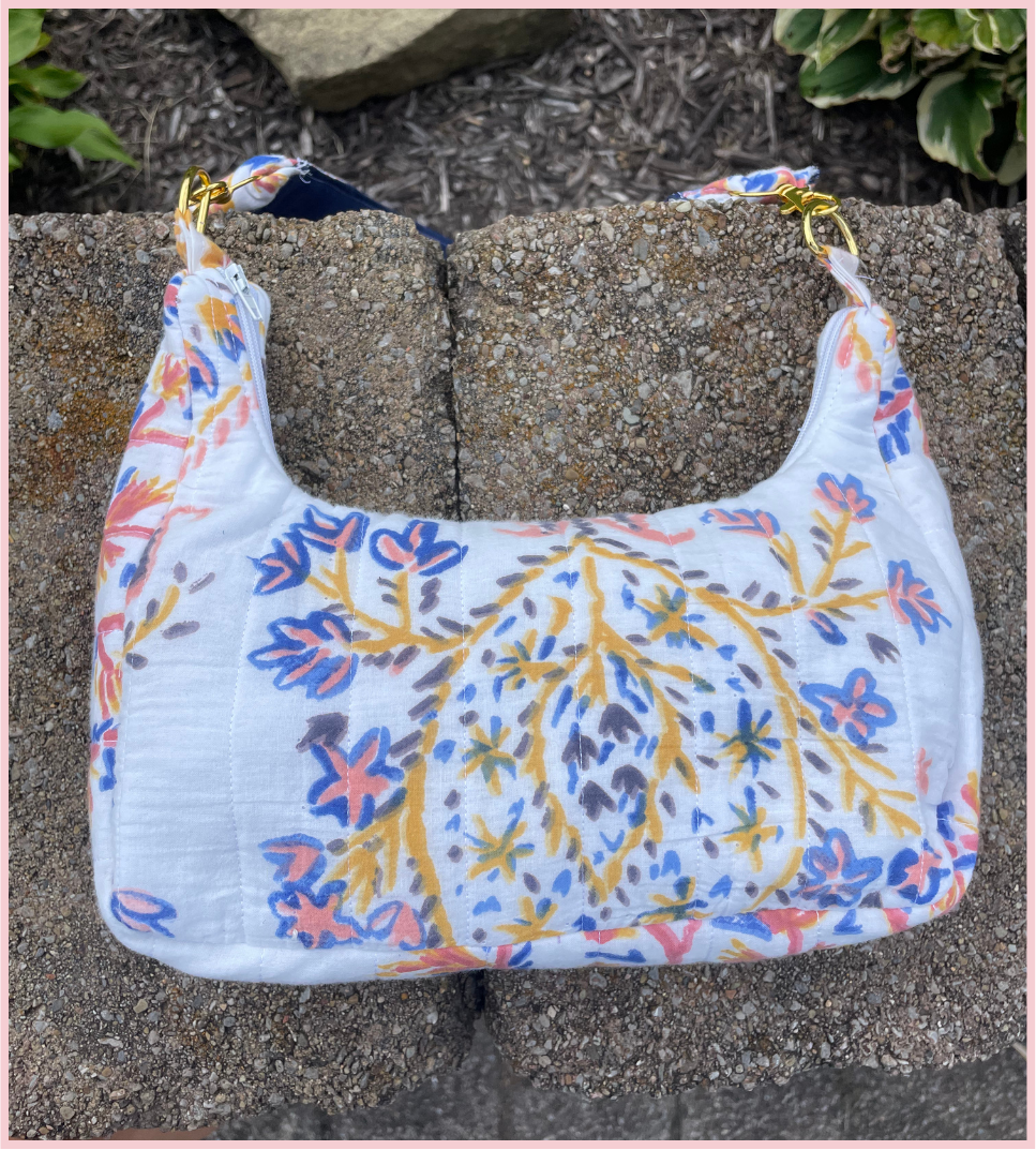 A white shoulder bag with a colorful floral and branch pattern in blue, pink, and yellow, resting on a stone wall outdoors.
