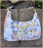 A white shoulder bag with a colorful floral and branch pattern in blue, pink, and yellow, resting on a stone wall outdoors.
