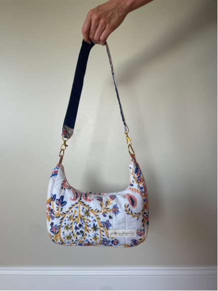 A hand holding a quilted shoulder bag with a white background and a colorful floral paisley pattern in blue, pink, and yellow. The bag has a dark navy blue adjustable shoulder strap with gold-tone hardware.