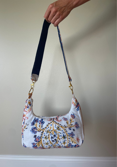 A hand holds up a small shoulder bag with a white base and a multicolor floral pattern in blue, orange, and yellow. The bag has a dark navy blue adjustable shoulder strap with gold-tone hardware.