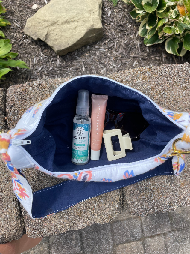 A white canvas zippered handbag with a colorful floral print and navy blue interior, shown open on a stone surface with various toiletries inside.