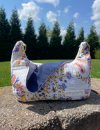 A small fabric shoulder bag with a floral print and a solid navy blue accent panel, sitting on a concrete ledge outdoors with grass, trees, and a blue sky in the background.