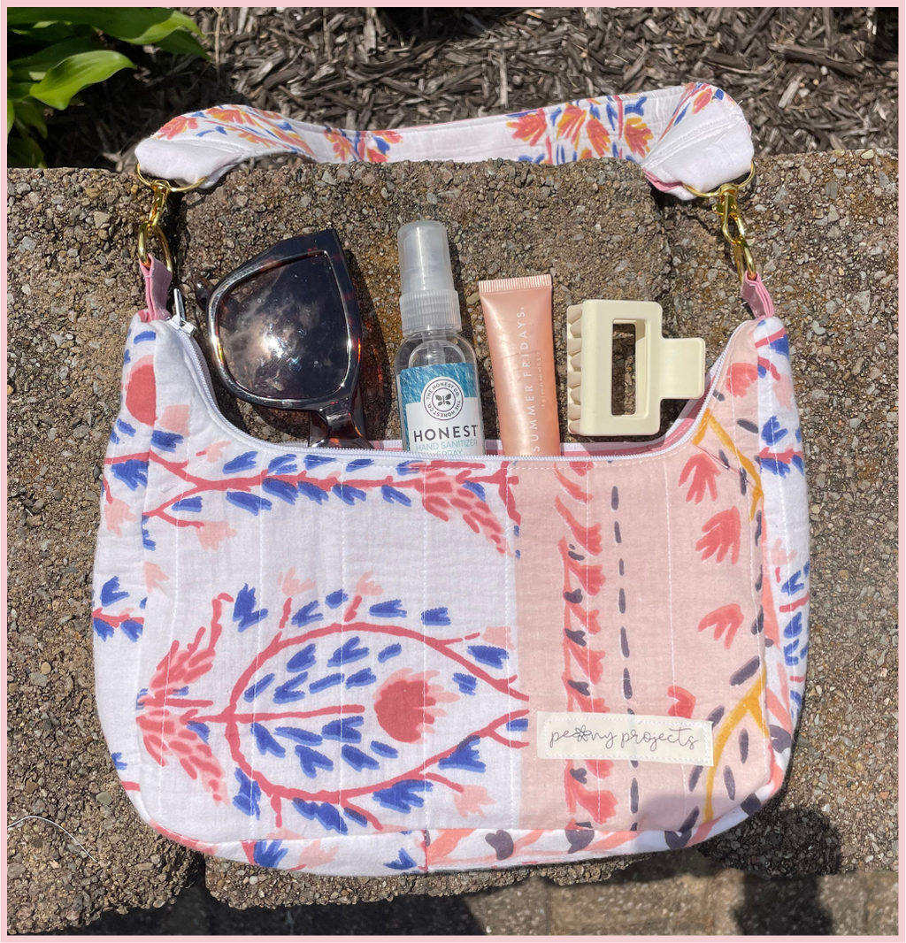A shoulder bag with a white base and a colorful botanical patchwork print in pink, blue, and coral, shown outdoors on a stone surface with sunglasses, a hand sanitizer bottle, a lip balm tube, and a hair clip inside.