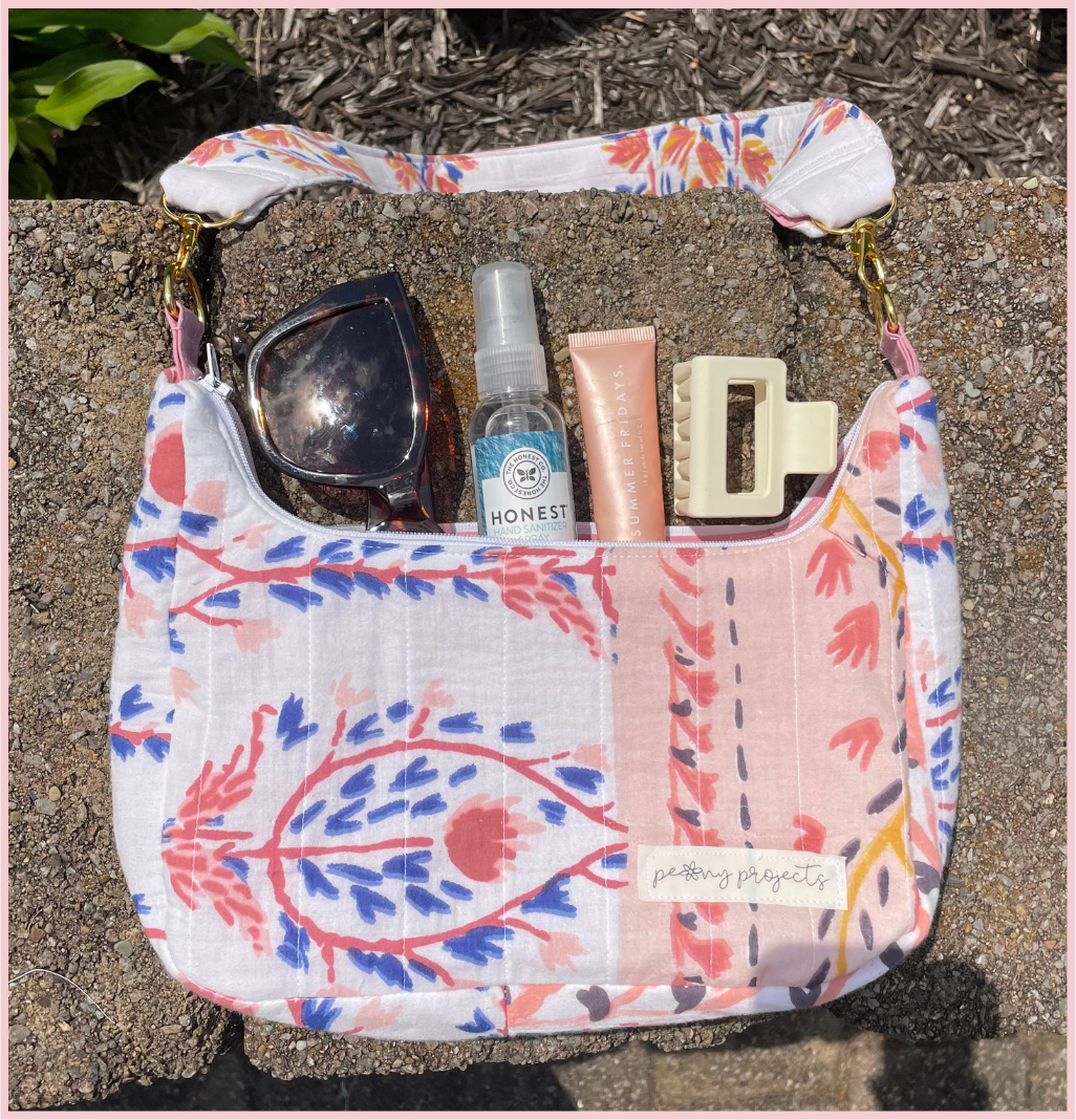 A shoulder bag with a white base and a colorful botanical patchwork print in pink, blue, and coral, shown outdoors on a stone surface with sunglasses, a hand sanitizer bottle, a lip balm tube, and a hair clip inside.