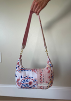A handmade patchwork fabric shoulder bag with a floral and botanical print in pink, blue, and red on a white background, held by a hand against a beige wall.