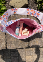 A floral shoulder bag with a pink interior is shown open on stone pavers outdoors, containing various travel items including a bottle, tube, sunglasses, and a hair clip.