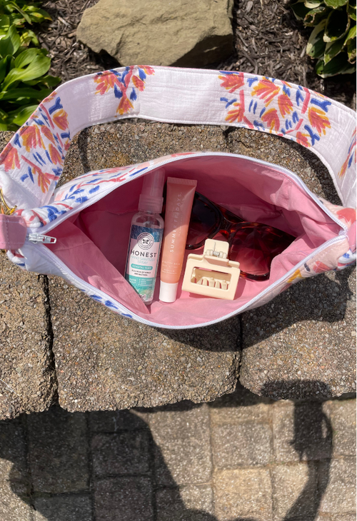 A floral shoulder bag with a pink interior is shown open on stone pavers outdoors, containing various travel items including a bottle, tube, sunglasses, and a hair clip.