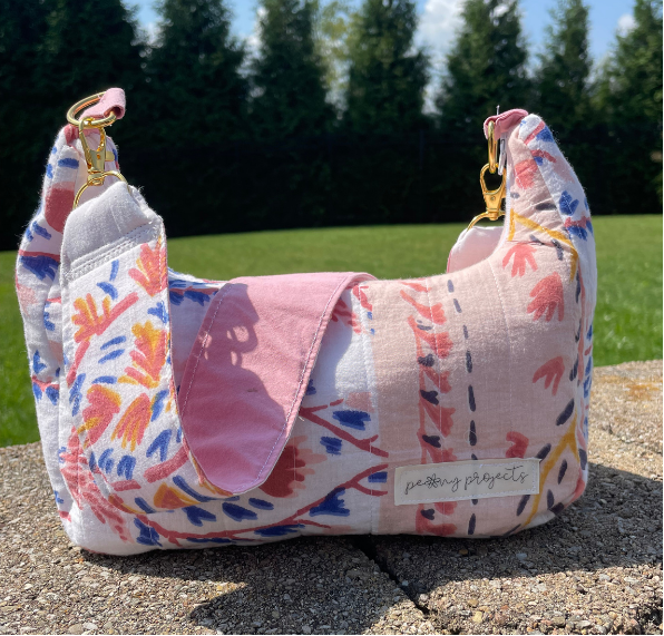 A handmade shoulder bag made from mixed fabric scraps, featuring a pink flap, a multicolor floral and geometric print body, and gold-tone hardware, sitting outdoors on a stone surface with a green lawn and trees in the background.