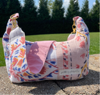 A handmade shoulder bag made from mixed fabric scraps, featuring a pink flap, a multicolor floral and geometric print body, and gold-tone hardware, sitting outdoors on a stone surface with a green lawn and trees in the background.