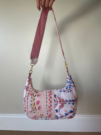 A handmade shoulder bag made from pink, blue, and white patterned fabric panels, held up by a hand against a beige wall.