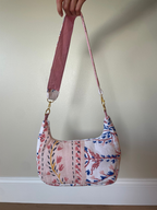 A handmade shoulder bag made from pink, blue, and white patterned fabric panels, held up by a hand against a beige wall.