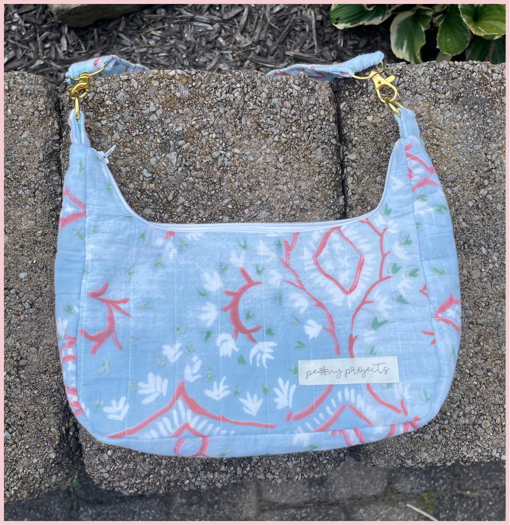 A light blue fabric shoulder bag with a coral and white botanical pattern, resting on a concrete surface. The bag features a white label reading 'putting plants to sleep' and gold-tone hardware.