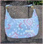 A light blue fabric shoulder bag with a coral and white botanical pattern, resting on a concrete surface. The bag features a white label reading 'putting plants to sleep' and gold-tone hardware.