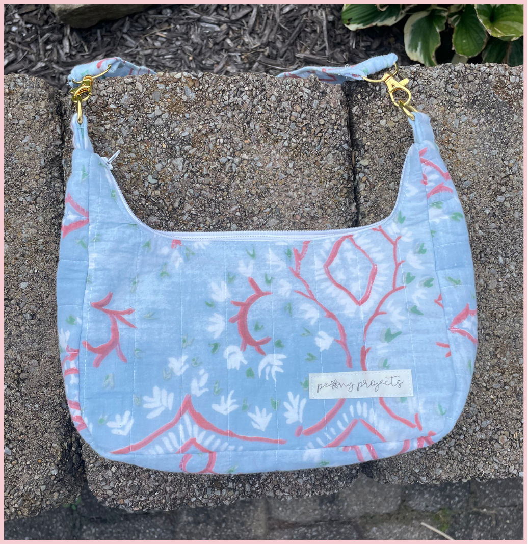 A light blue fabric shoulder bag with a coral and white botanical pattern, resting on a concrete surface. The bag features a white label reading 'putting plants to sleep' and gold-tone hardware.