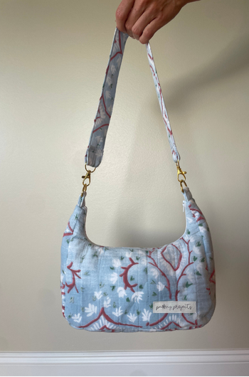 A light blue fabric shoulder bag with a red and white floral branch pattern, held by a hand against a beige wall.