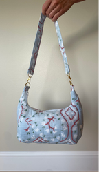 A light blue quilted fabric shoulder bag with a red and green floral vine pattern, held by a hand against a beige wall.