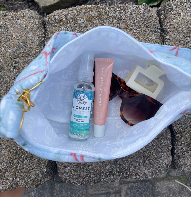 A light blue and white patterned drawstring pouch filled with travel items, including a bottle of Honest Anti-Aging Facial Mist, a pink tube of Summer Fridays body cream, tortoiseshell sunglasses, and a cream hair clipper, set on stone pavers.
