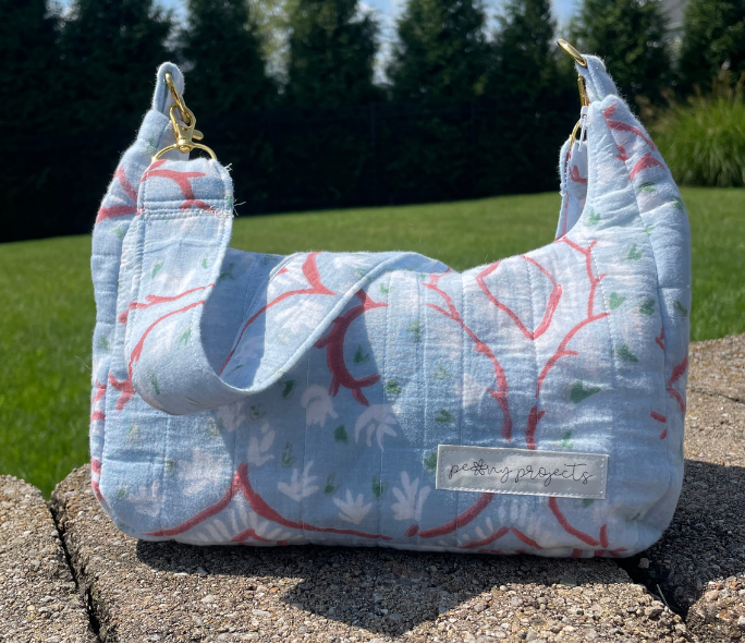 A quilted fabric shoulder bag with a light blue background and a red and white floral pattern, resting on a stone surface outdoors with a green lawn and trees in the background. A brand label reading 'petite projects' is visible on the front.