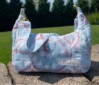 A quilted fabric shoulder bag with a light blue background and a red and white floral pattern, resting on a stone surface outdoors with a green lawn and trees in the background. A brand label reading 'petite projects' is visible on the front.